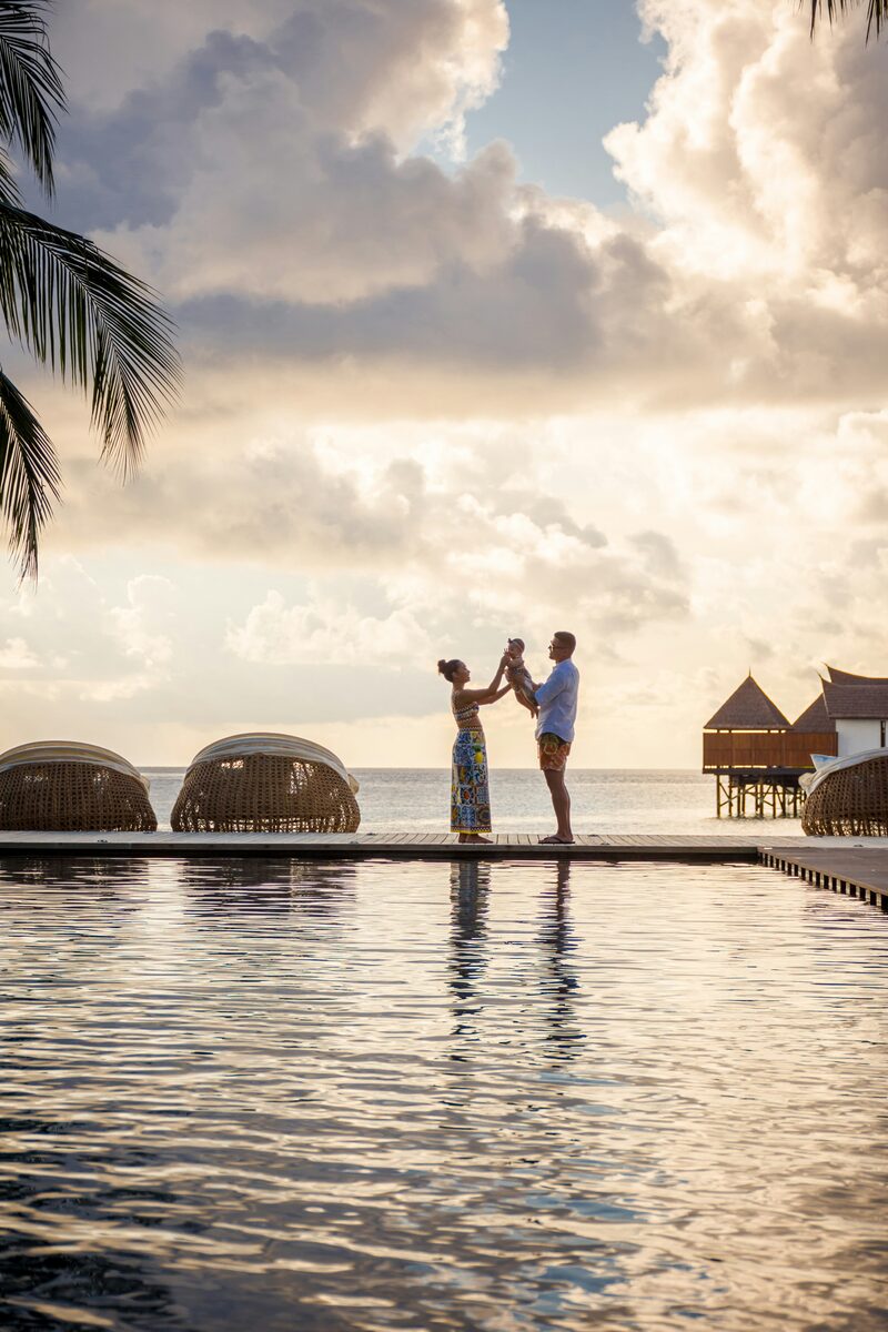 Parents holding their baby on an overwater resort dock at sunset